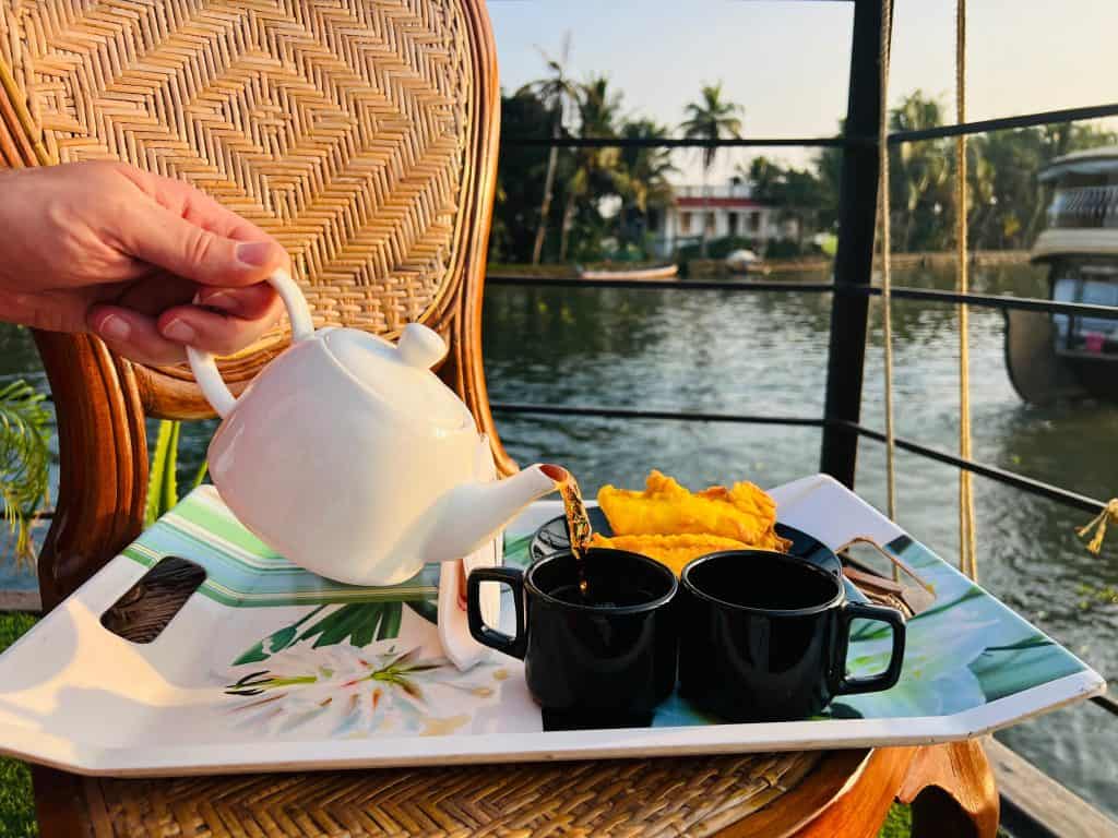 Beautiful Kerala backwaters: white teapot with masala chai being poured and 2 black tea cup. A plate of banana fry and views of the backwaters in the background.