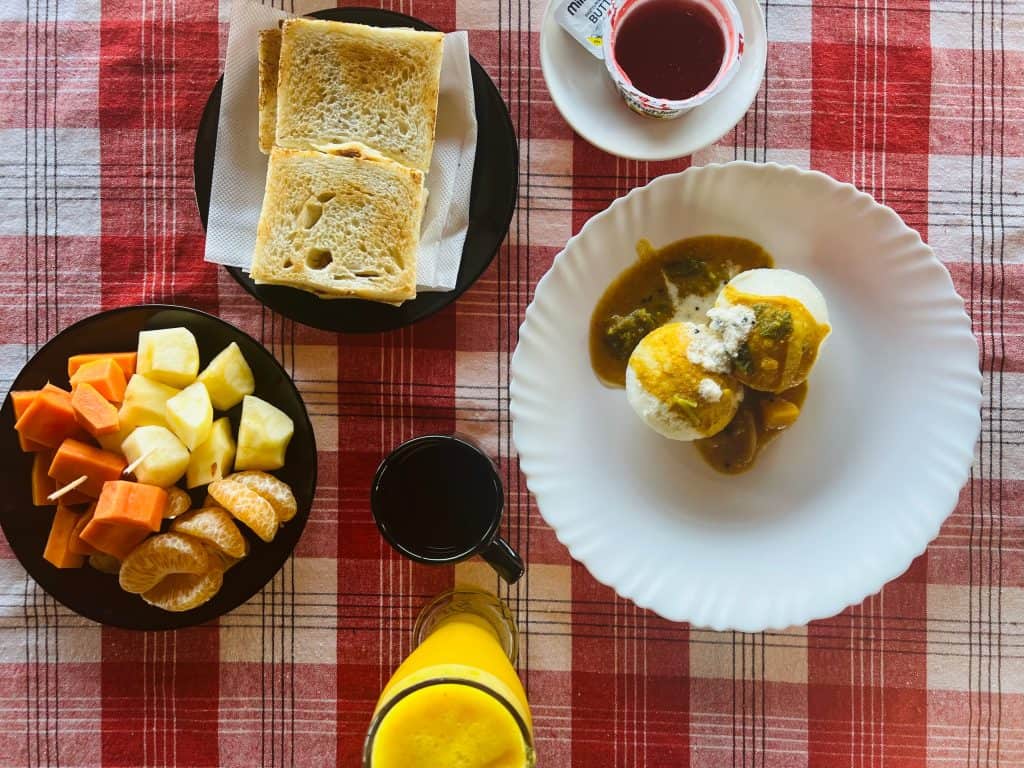 Flat lay photograph of breakfast on the houseboat. Toast, idli sambhar, fruits, ornage juice and coffee on a red checked table cloth,