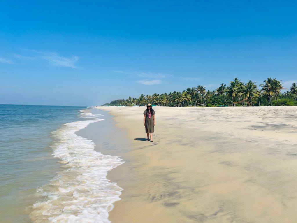 Marari Beach travel guide: bejal walking down the beach with golden sand and the waves coming in, with palm trees swaying in the background. Bejal is wearing a khaki dress, Fedora hat with a pink flower necklace