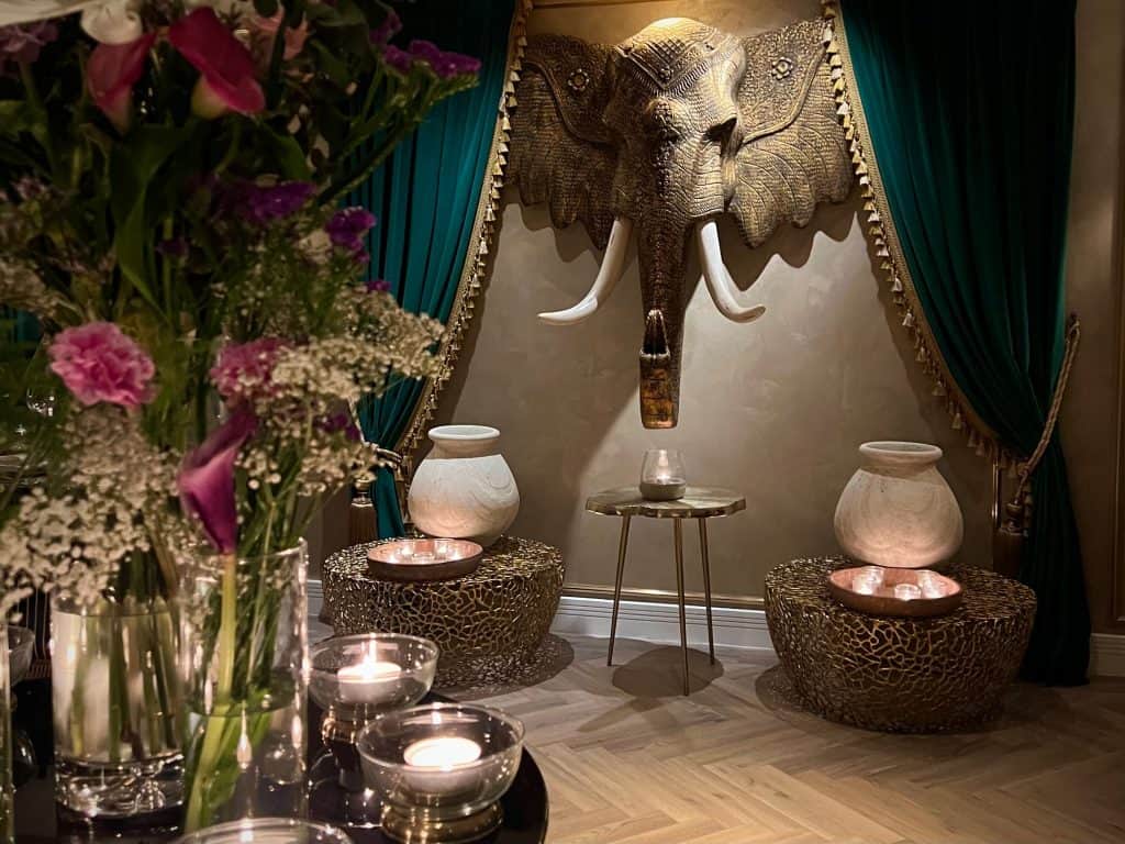 Indian Fine Dining At Varanasi.  A zoomed out image of the bronze elephant head with a table of flickering candles and lanterns in front. There is also a fresh bouquet of flowers here at Varanasi, Leicester