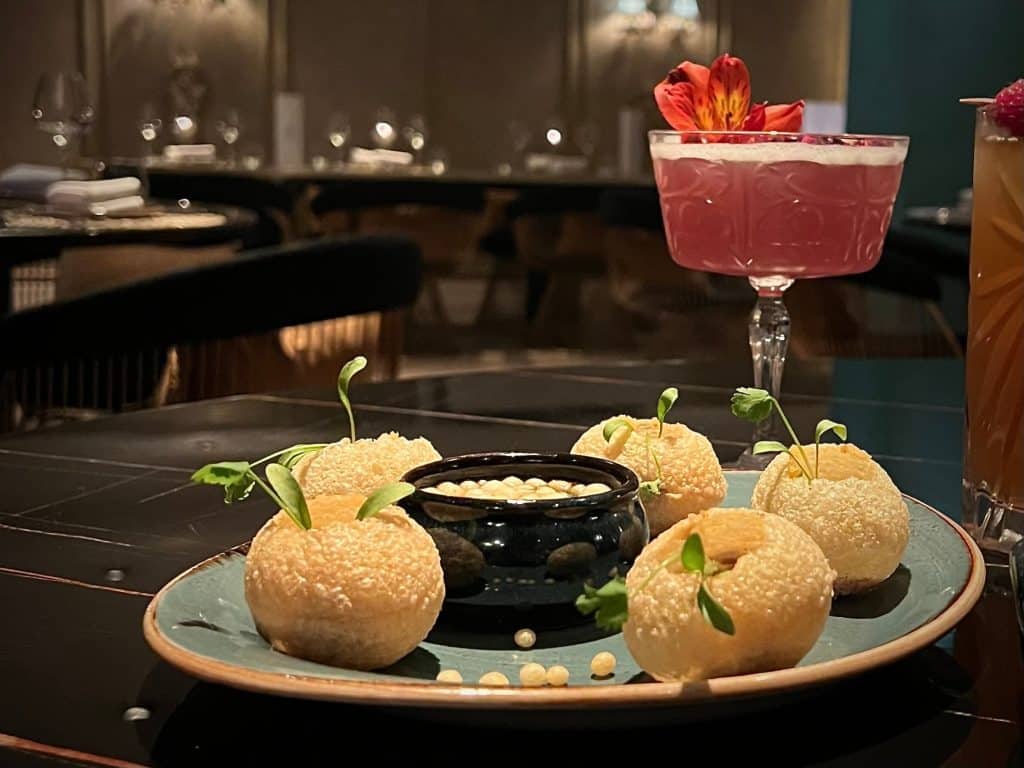 5 puffed pani puri on a plate with mint and coriander water and in the background there's a red/pink lavender rose garden martini with atmospheric lighting. Location is Varanasi, Leicester