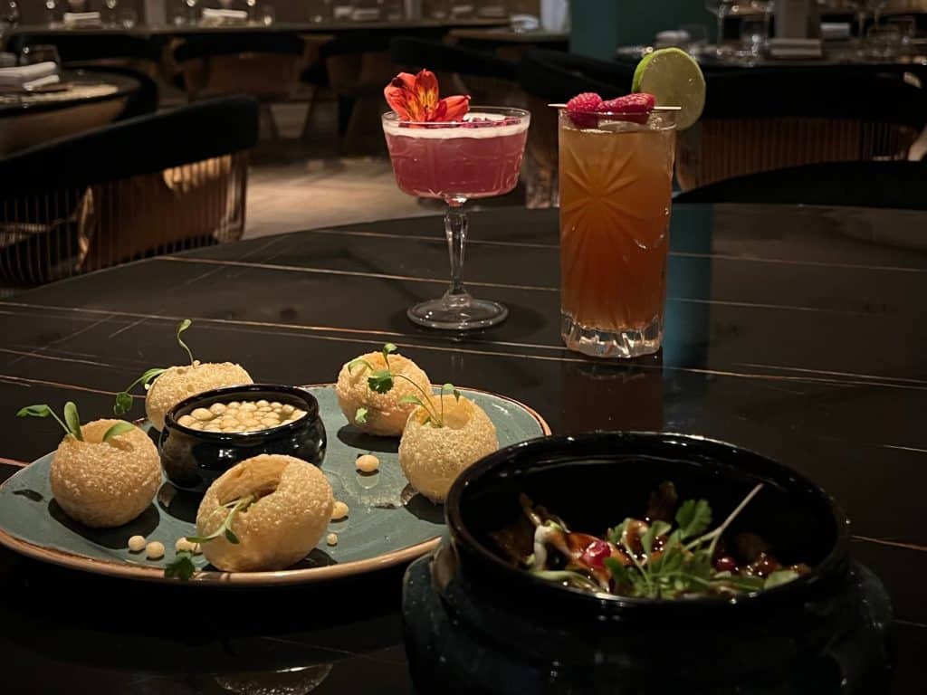 pani puri with coriander and mint water on a black plate with the papri chat at the side. in the background is the dark and stormy and lavender garden martini cockatils.