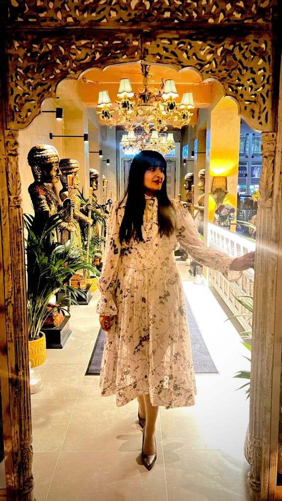 Bejal standing near entrance of Varanasi wearing a floaty white dress and grey pointed heels.