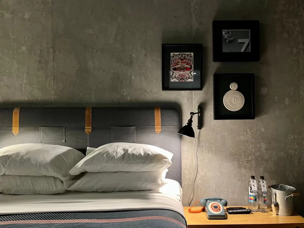 Moxy queen bedroom in San Diego with pillows stacked and side light plus the quirky telephone. There are grey walls and white pillows plus bright orange splashes of colour with the decor.