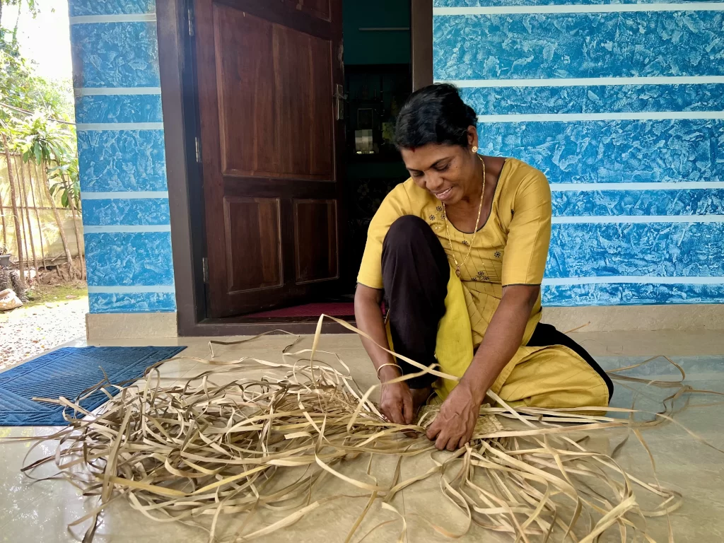 Lady weaving mats at Viakom village tour. She is wearing a long yellow top and black trousers and is sitting on the floor of her veranda.