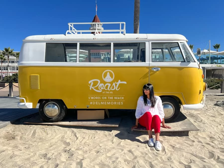 Bejal wearing red trousers and white top sitting outside the yellow Roast at The Del campervan