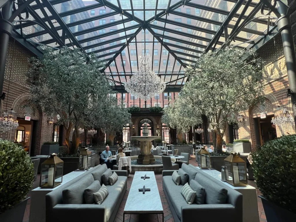 The inner grand courtyard of the 3 Arts Club Cafe at RH Chicago. The space has a glass ceiling with chandelier. There are grey sofas at the front with table seating all around the room with a fountain water feature in the centre.