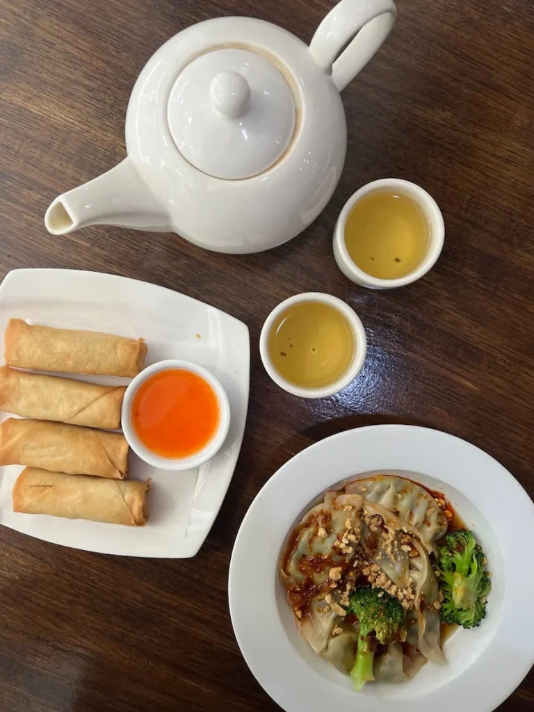 A flat lay photo of a teapot with 2 cups of jasmine tea, a plate of spring rolls and dipping sauce and spicy dumplings with chilli sauce at Veggie House, Chicago