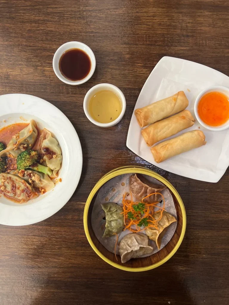 A flat lay photo of a teapot with 2 cups of jasmine tea, a plate of spring rolls and dipping sauce and spicy dumplings with chilli sauce. plus vegetable dumplings at Veggie House, Chicago