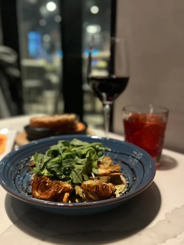 The artichoke salad on a charcoal plate with glaze and a red wine in the background with the red strawberry fields cocktail next to it at The Albert at Hotel EMC2. one of the places with vegetarian friendly food in Chicago