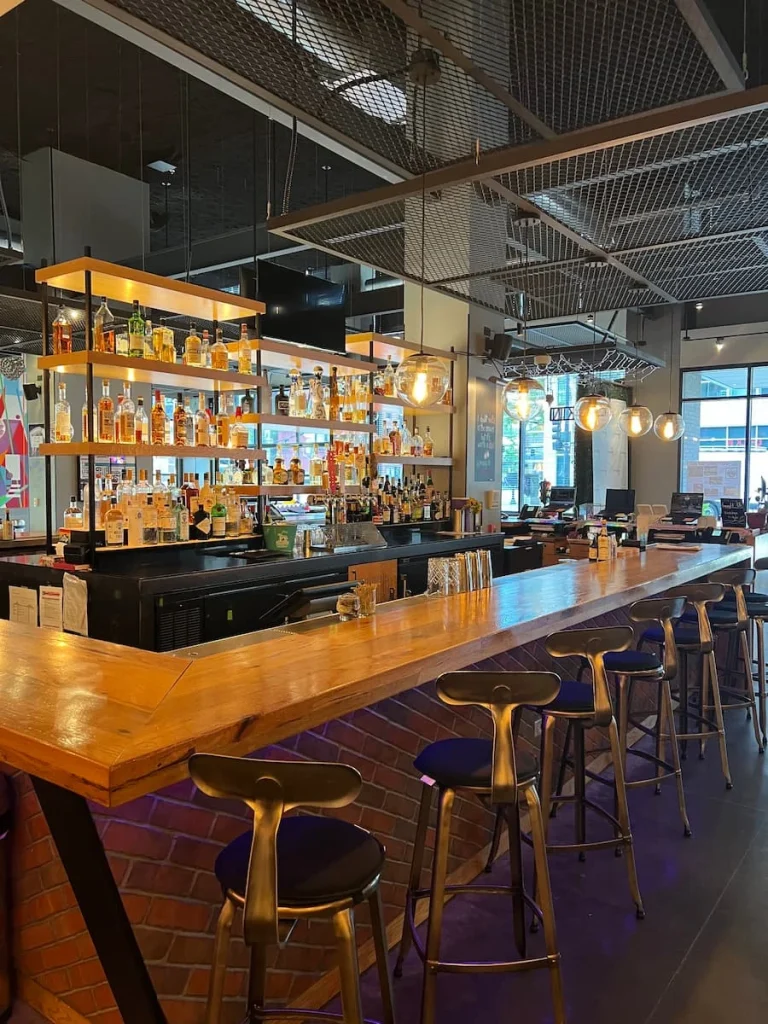 Bar area at Moxy, Chicago. Spirits displayed on the glass shelves, with high top stools and a tan shiny bar top