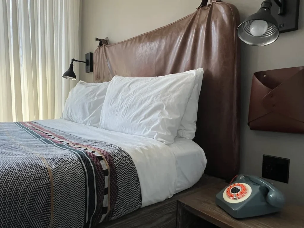 Bed and bedside table at Moxy, Chicago featuring retro grey and orange phone, white sheets and gry and orange bed spread, plus a faux leather tan headboard.