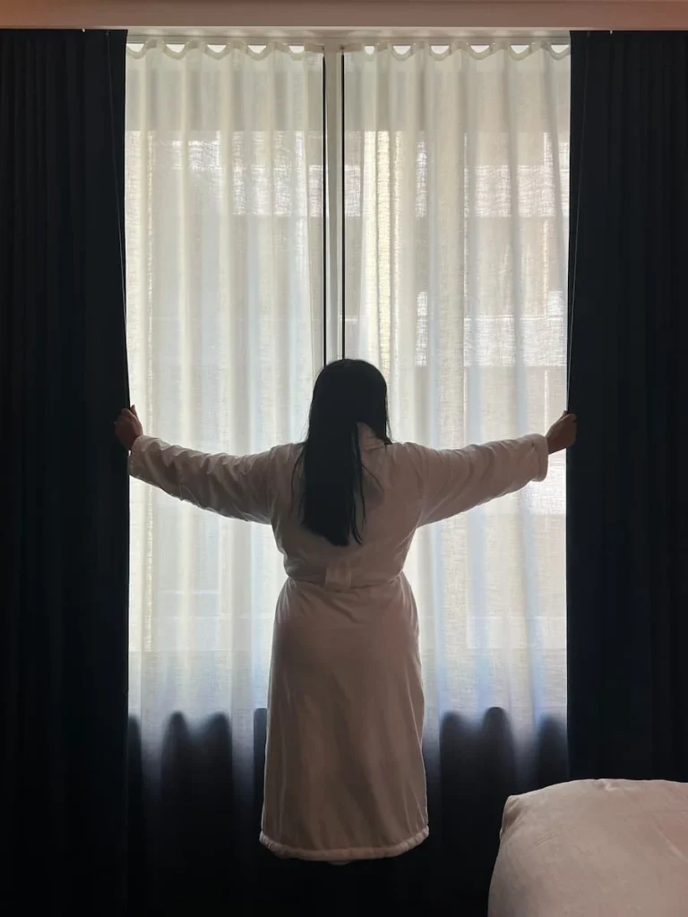 Bejal drawing the curtains back wearing a dressing gown in the queen room at Hotel EMC2, one of the cool and quirky boutique hotels in Chicago