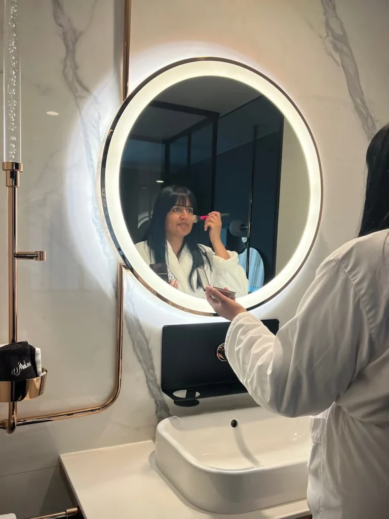 Bejal putting on foundation wearing a white dressing with a reflection in the light rimmed mirror in the queen room at Hotel EMC2, one of the cool and quirky boutique hotels in Chicago