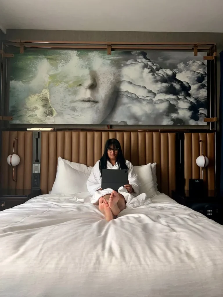 Bejal sitting on the Queen Room bed in a dressing gown working on ipad infront of a gold hued padded headboard above which there is a sci-fi artwork of a woman's face at Hotel EMC2, one of the cool and quirky boutique hotels in Chicago