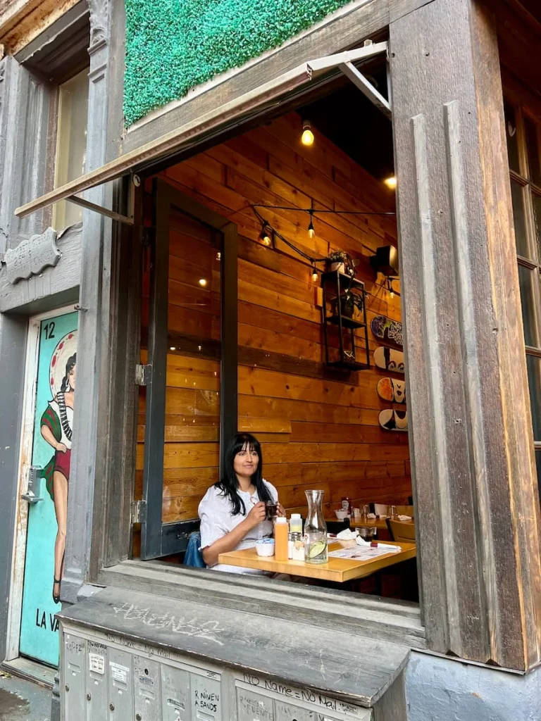 Bejal sitting in window seat of Frida Room, Chicago wearing a cream and white floral dress looking out of the window onto the street in Pilsen