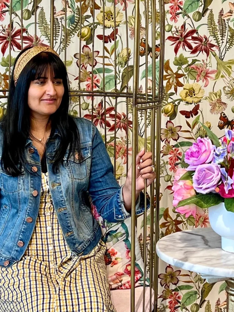 Bejal sitting on gold cage looking at flowers at Sugary, Chicago. bejal is wearing a blue, white and yellow checked dress, denim jacket and straw hairband. She is looking at a bunch of artificial white and pink peony's and the background has a floral statement wall paper with small butterfly print.
