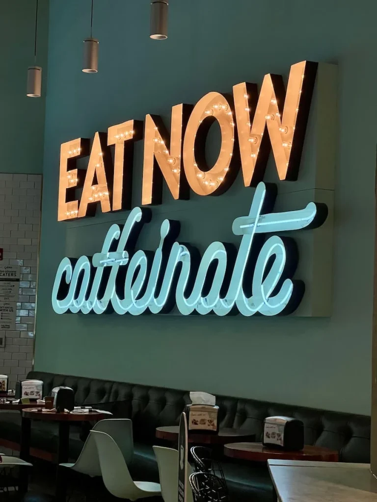 Eat Now Coffee sign at Goddess & Baker, Streeterville, Chicago on a teal background with text in orange and blue with a light outline.