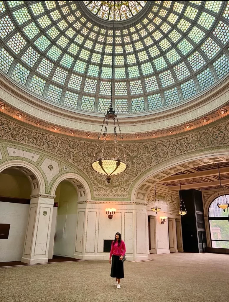 World's largest Tiffany Dome with mint blues and gold and a light piece hanging down. bejal stands in the middle of the room looking at the dome wearing a bright pink jumper and black skirt