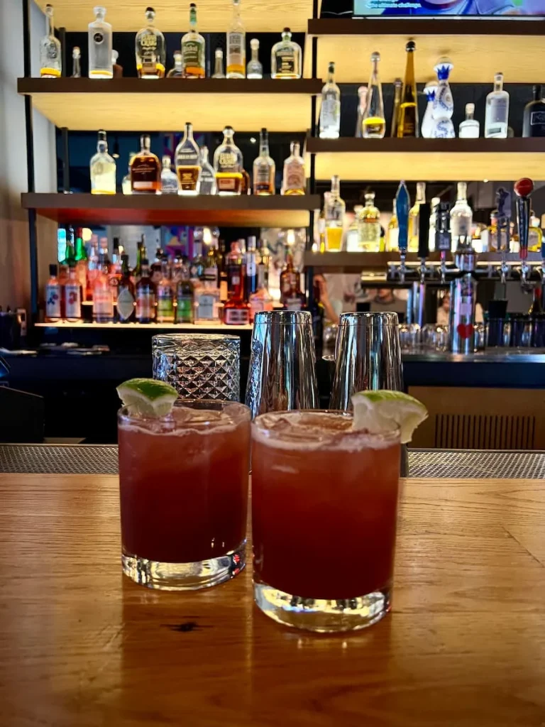 GOT Moxy welcome cocktail at Moxy, Chicago. 2 dark red cocktails on teh bar top with ice and a slice of lime on the rim with the bar back shelve of spirits in the background