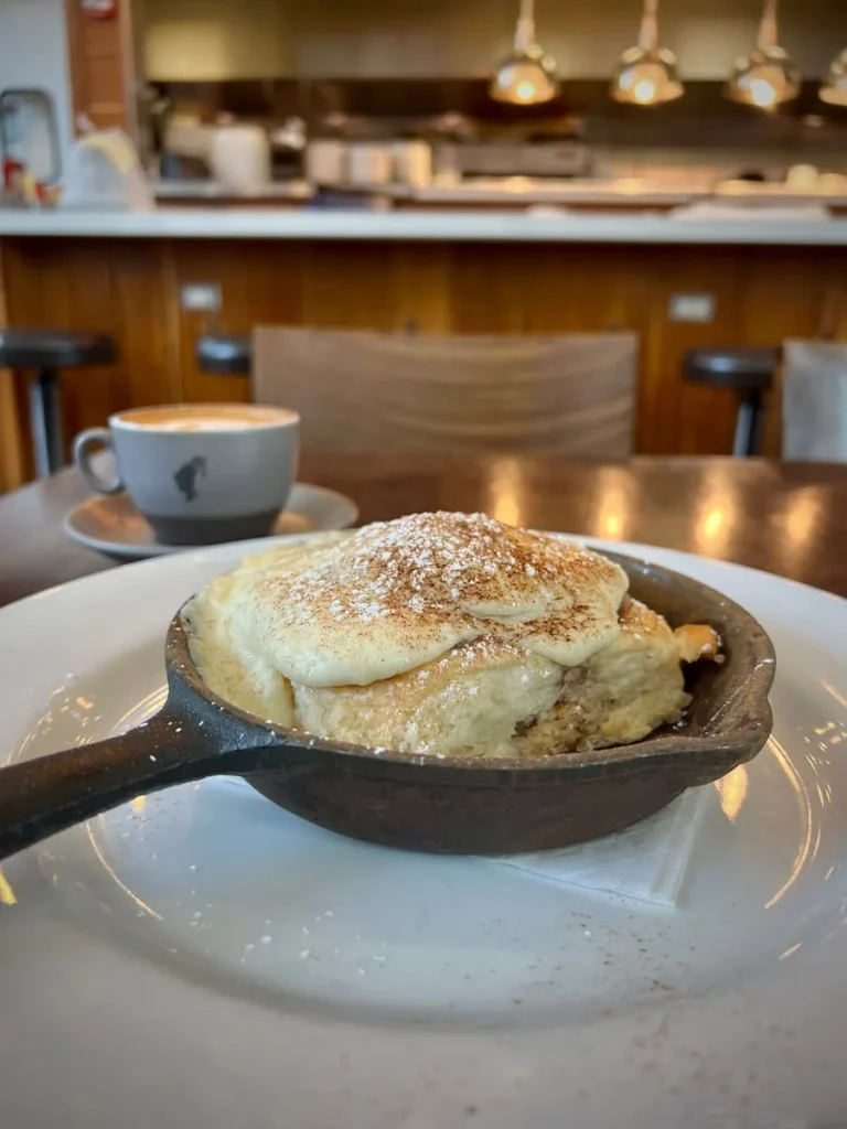 Glazed cinnamon roll topped with cream chese frosting and sugar in a skillet with the bar and cappuccino in the background at Kanela Breakfast Club, Chicago