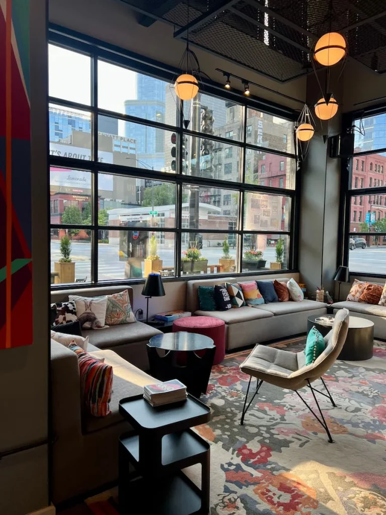 Lounge area at Moxy, Chicago, with a grey sofa and multicolour cushions. A view of the street through the large floor to ceiling warehouse style windows.