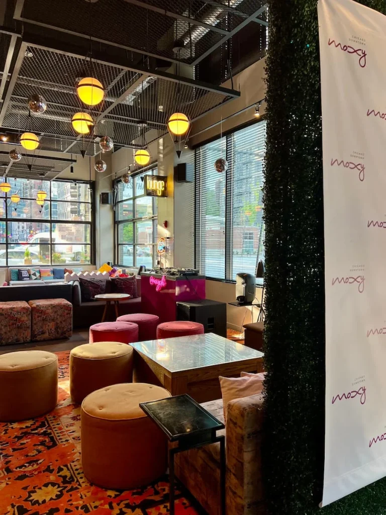 Lounge spaces with DJ decks at Moxy Chicago. the multicolour interiors and floor to ceiling windows and deep pink cushioned pouffes fill the excess space.