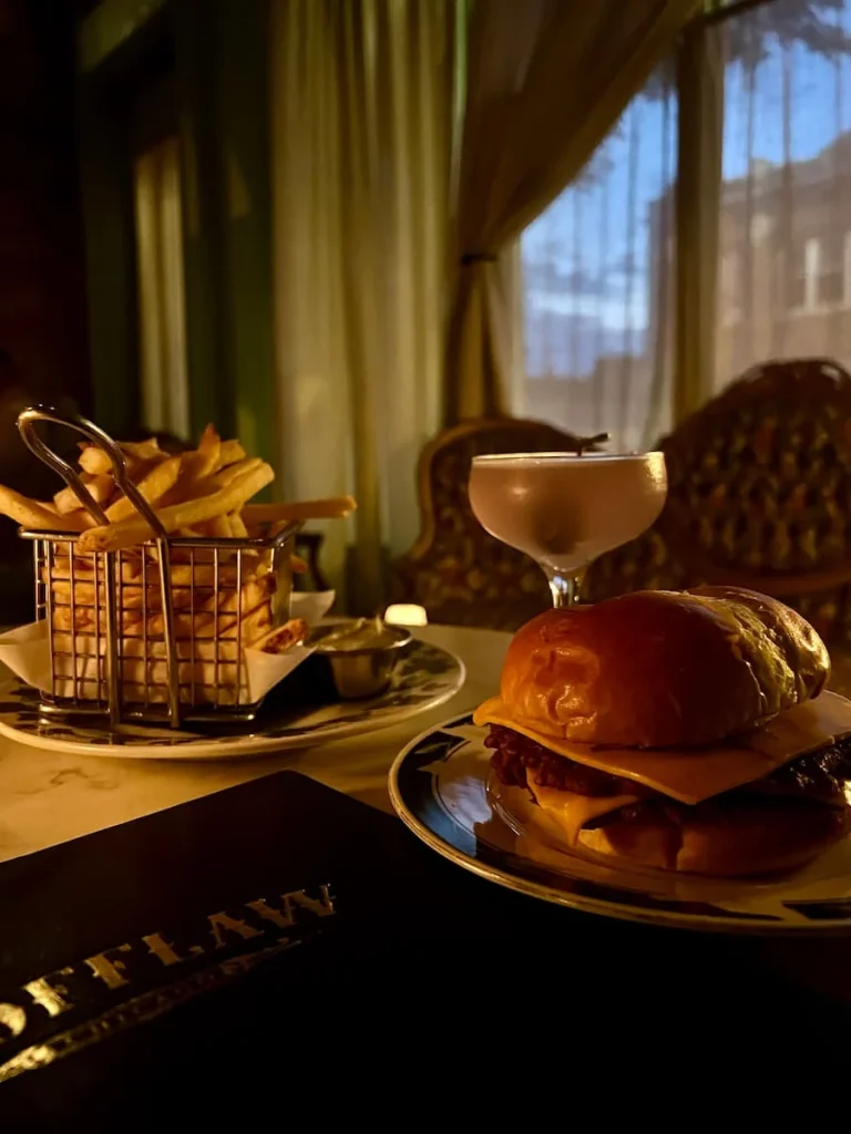 Plant-based burger, Another Plane cocktail and chips at Scofflaw, Chicago. The background is dark with a sofa and chandelier in the background