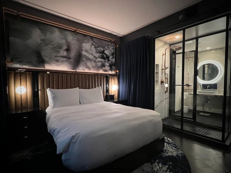 Queen bedroom with wide angle side shot of white linen on bed, featuring artwork above tthe headboard and the open shower cubicle showing to the side at Hotel EMC2, one of the cool and quirky boutique hotels in Chicago