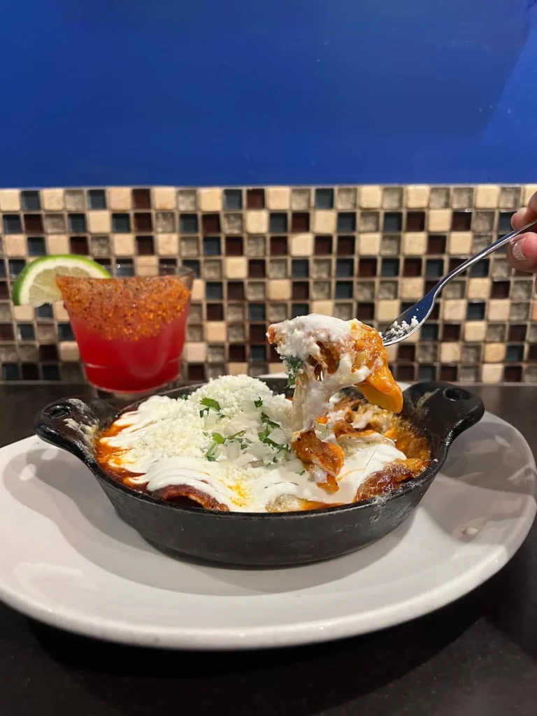 Roasted Vegetable enchiladas at XOCO, Chicago in a skillet pan with a fork of enchiladas being lifted up. The back wall has brown and white mosaic tiles and a bright blue wall.