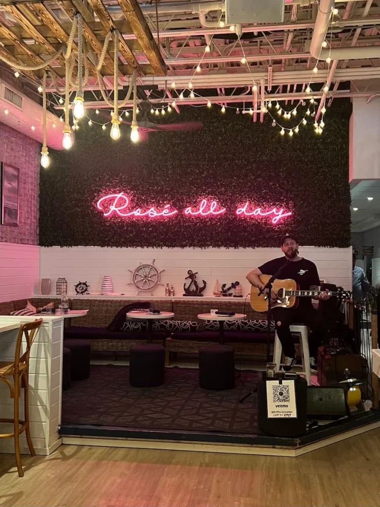 Rose all day, pink neon sign at The Hampton Social Club in Chicago. A musician is playing a guitar on the podium in front of the sign.