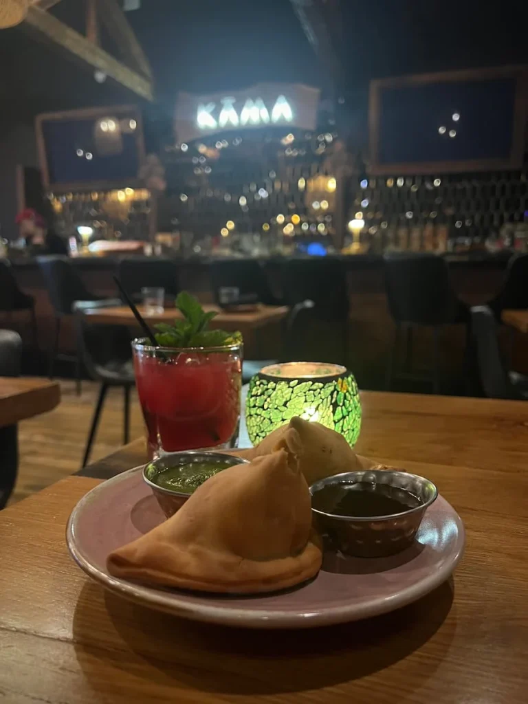 vegetable Samosas and watermelon smash cocktail side view at Kama, Chicago. There is a green mosaic glass table lamp and teh samosa are served with a green coriander chutney and dark brown tamarind chutney