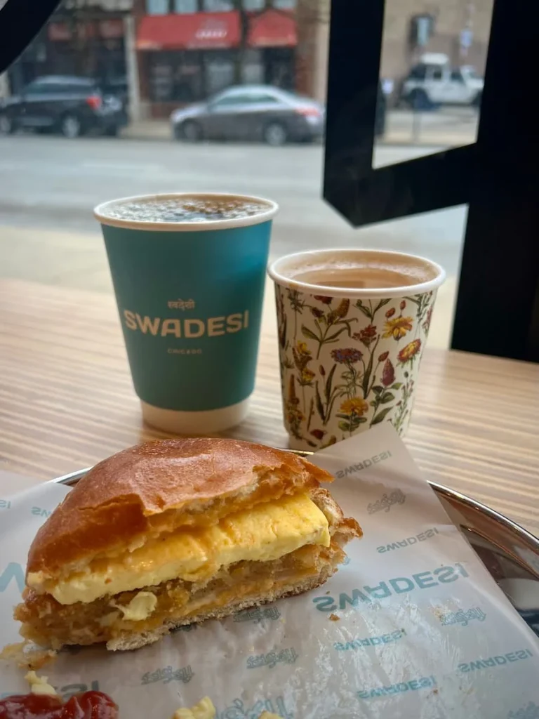 Side view of vegetarian breakfast sandwich and masala chai in a flower paper cut and a black coffee in a blue paper cup at Swadesi, Chicago. One of the places to eat vegetarian-friendly food in Chicago