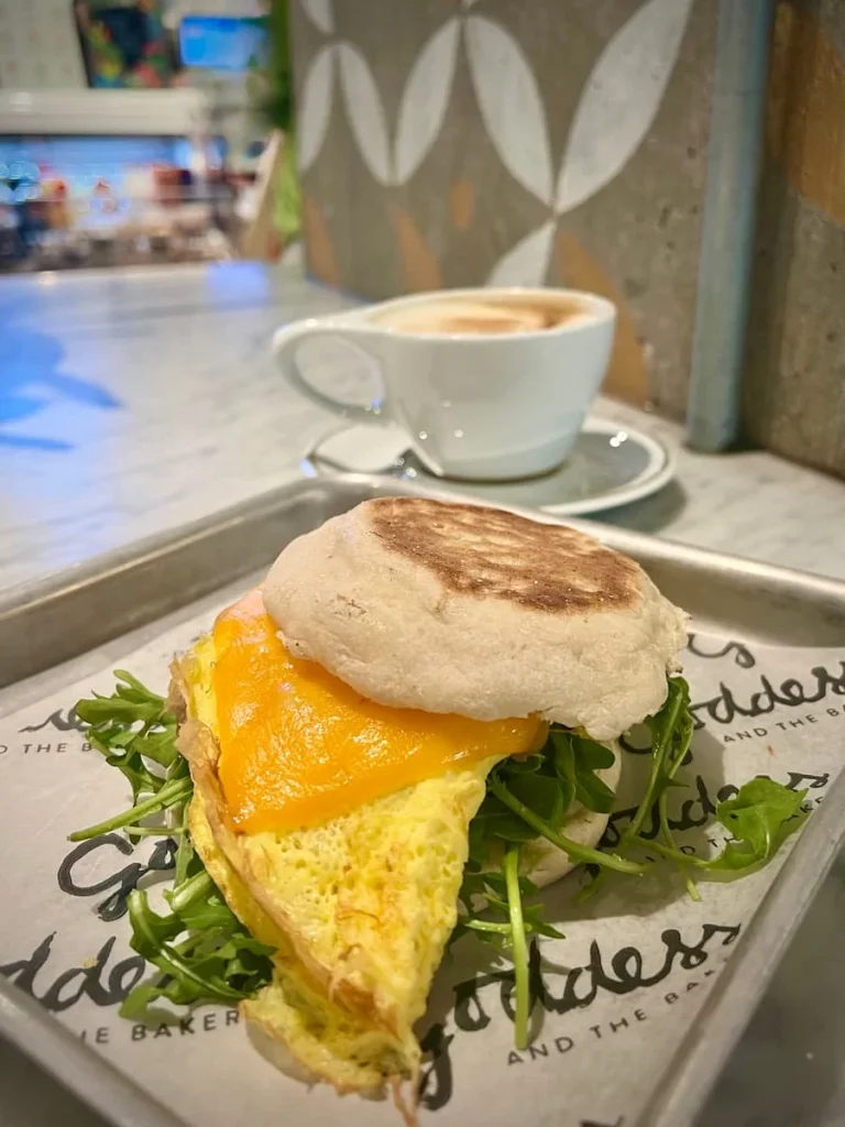 The impossible Breakfast muffin with based sausage, omelette, arugula, avocado and more with a cappuccino in the background at Goddess & Baker, Streeterville, Chicago