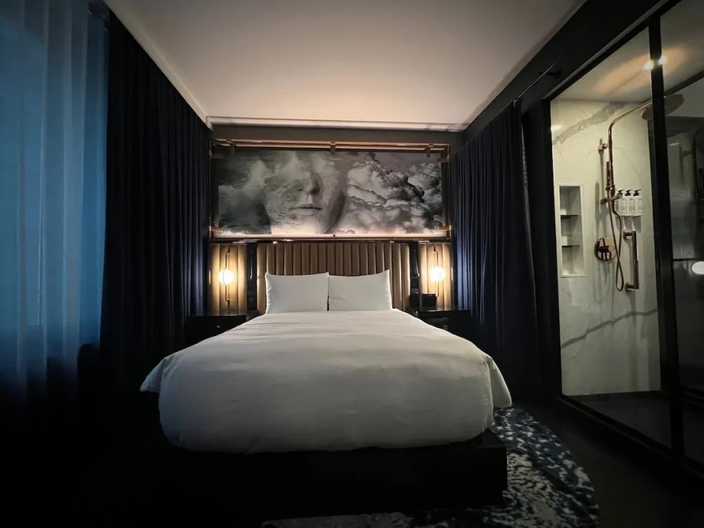 A wide angle shot of the Queen Room at Hotel EMC2 featuring the bed, over bed, artwork and partial glass cube open shower at Hotel EMC2, one of the cool and quirky boutique hotels in Chicago