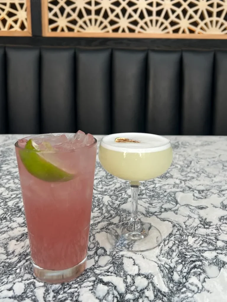 Cocktails on a grey and white marble effect table. Cocktails are Berry Tequila or Mezcal Sour. There is a black cushioned portion in the background with gold design panelling. Location is Roof on thewit, Chicago