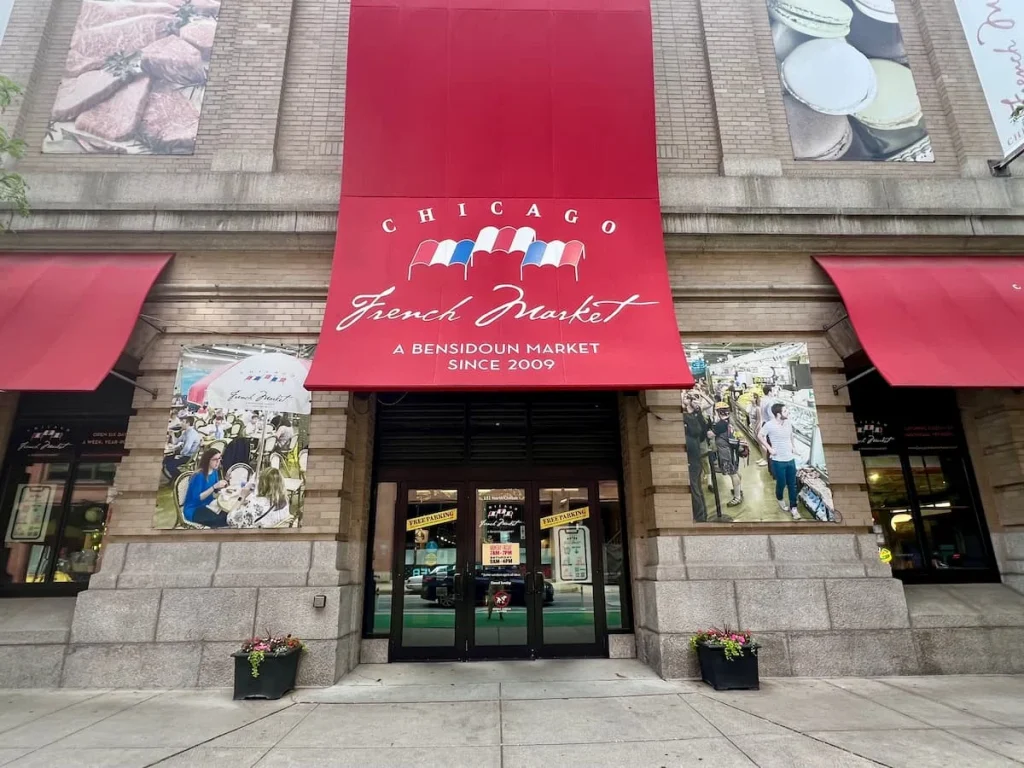 Exterior of the French Market in Chicago which has a red awning with 3 flags of France. There are also flower baskets on either side of the entrance.