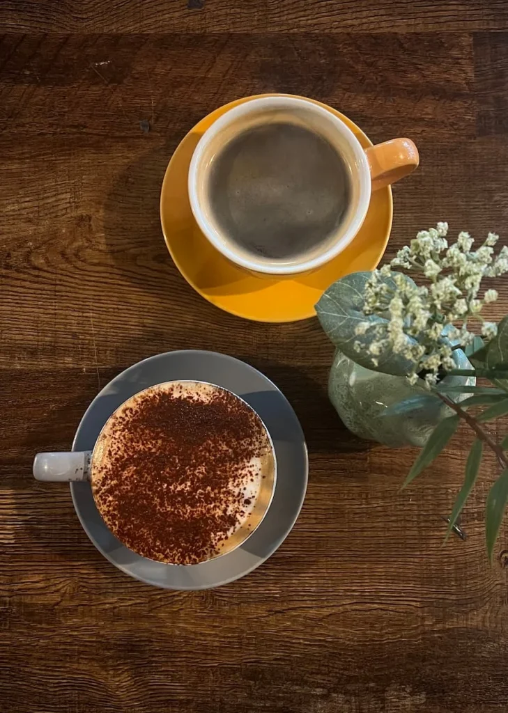 Black coffee and cappuccino flat lay at Lynn, Leicester. The image is a flat ay of a yellow coffee cup and saucer with a black coffee and a grey coffee cup and saucer with a cappuccino in it on a dark brown table top. There are some small jib flowers in a grey vase at the side