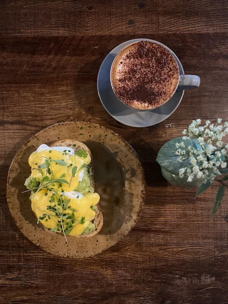 Smashed avocado with poached eggs and cappuccino at Lynn, Leicester. The image is a flat lay of a grey coffee cup and saucer with a cappuccino inside and a brown stoneware plate with sourdough topped with 2 poached eggs, Hollandaise sauce and micro herbs. There is a small grey vase with jib flowers and the table top is dark brown wood