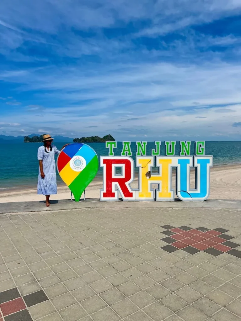 Things to do in Langkawi:  Tanjung Rhu beach sign with Bejal standing at the side in a blue and white checked dress