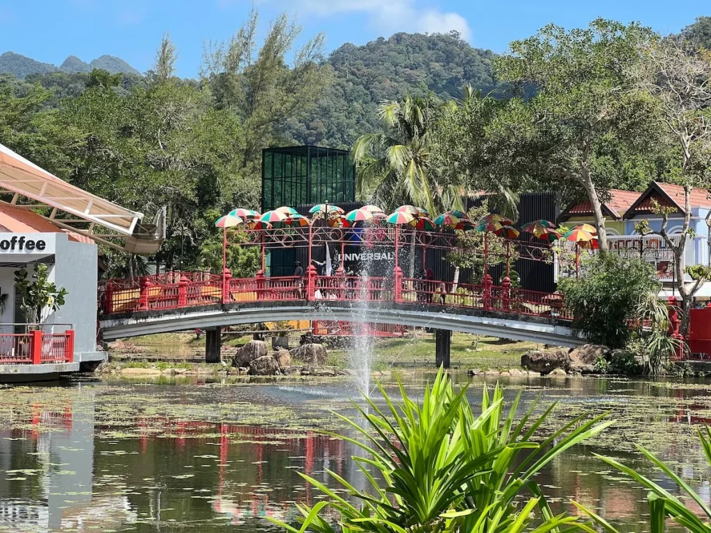 Things to do in Langkawi: Oriental Village bridge with fountain in front surrounded by greenery and colourful shops.