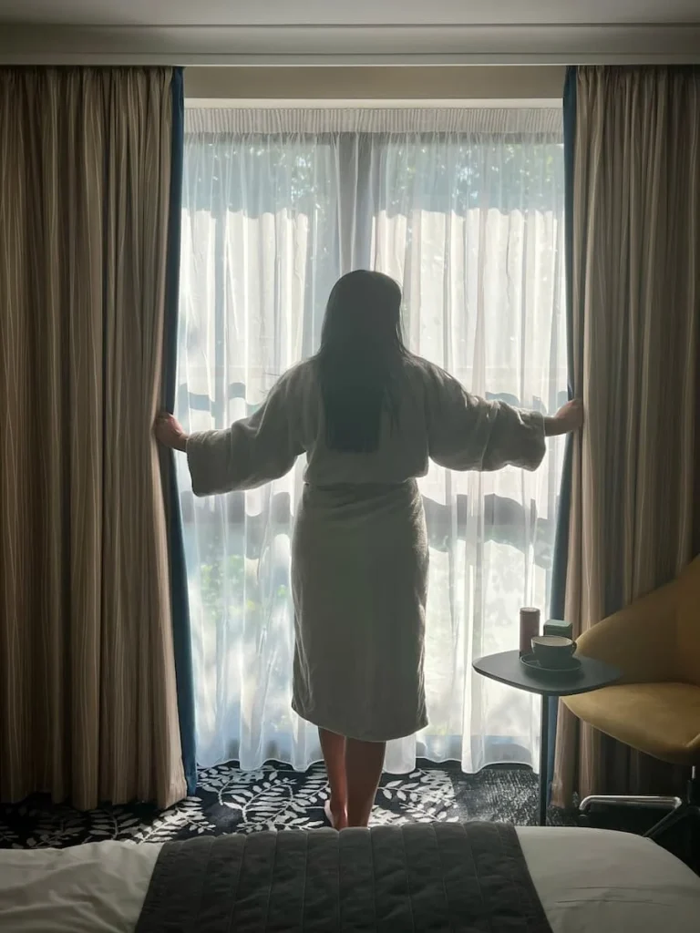 Bejal drawing the curtains in a bath robe at Burleigh Court Hotel, Loughborough