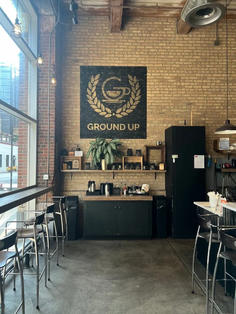 Interiors at Ground-up coffee on Erie street in Chicago. A counter for visitors on one side with high top stools and a 'Ground=Up' sign on the brick wall with a amenities station featuring milks, sugars, mixers, tissues and cup holders.