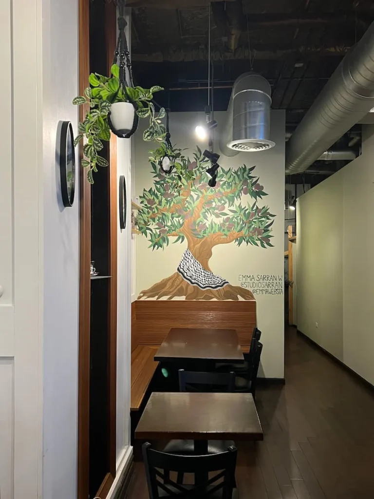 Interiors of the Oud coffee shop in Chicago. Tree artwork on the white walls with silver hanging lights, wooden tables and Palestinian inspired artifacts adorning the walls.