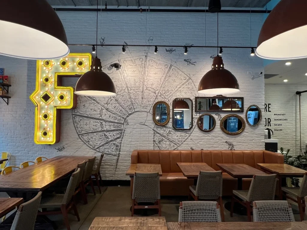 Mirror and cafe decor at Fairgrounds Coffee, Chicago. The back white wall has a fairground mural with different mirrors attached like a collage. There are also faux leather seats and bistro tables as well as cool dome lampshades in co-ordinating colours
