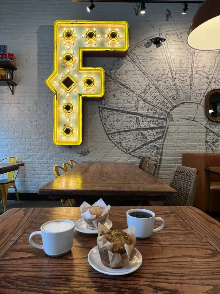 The big F letter with light bulbs inside and the Fairground mural on the white back wall. Infront on a brown topped table is a white mug od drip coffee and cappuccino with a plain muffin and limoncello muffin. 