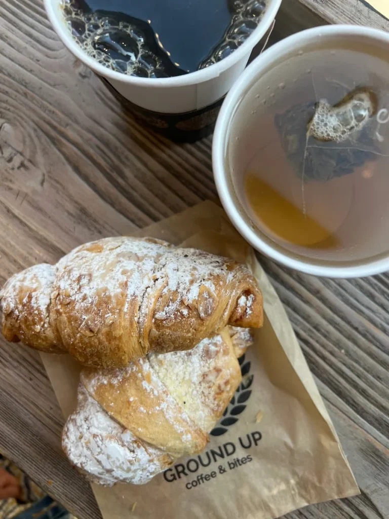 Top lay of almond croissants and hot drinks at Ground Up Coffee, Chicago. The hot drinks are black drip coffee and green jasmine tea. The 2 almond croissants are flakey adn dusted with icing sugar on a wooden top counter table with a 'Ground-Up' brown paper bag underneath.