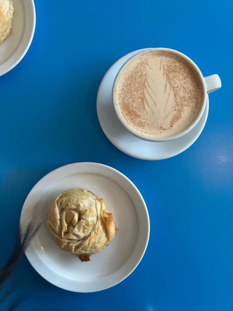 Top lay of a flaky Burek Croatian pastry next to a Chai Latte in a white cup and saucer, both on a bright blue table top surface at Doma Cafe, Chicago 