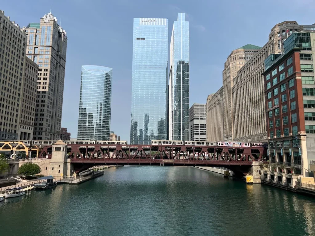 2 L trains going down Well's Street Bridge towards each other in Chicago. The image is captured with the skyscrapers in the background and river underneath on a sunny day with blue skies.