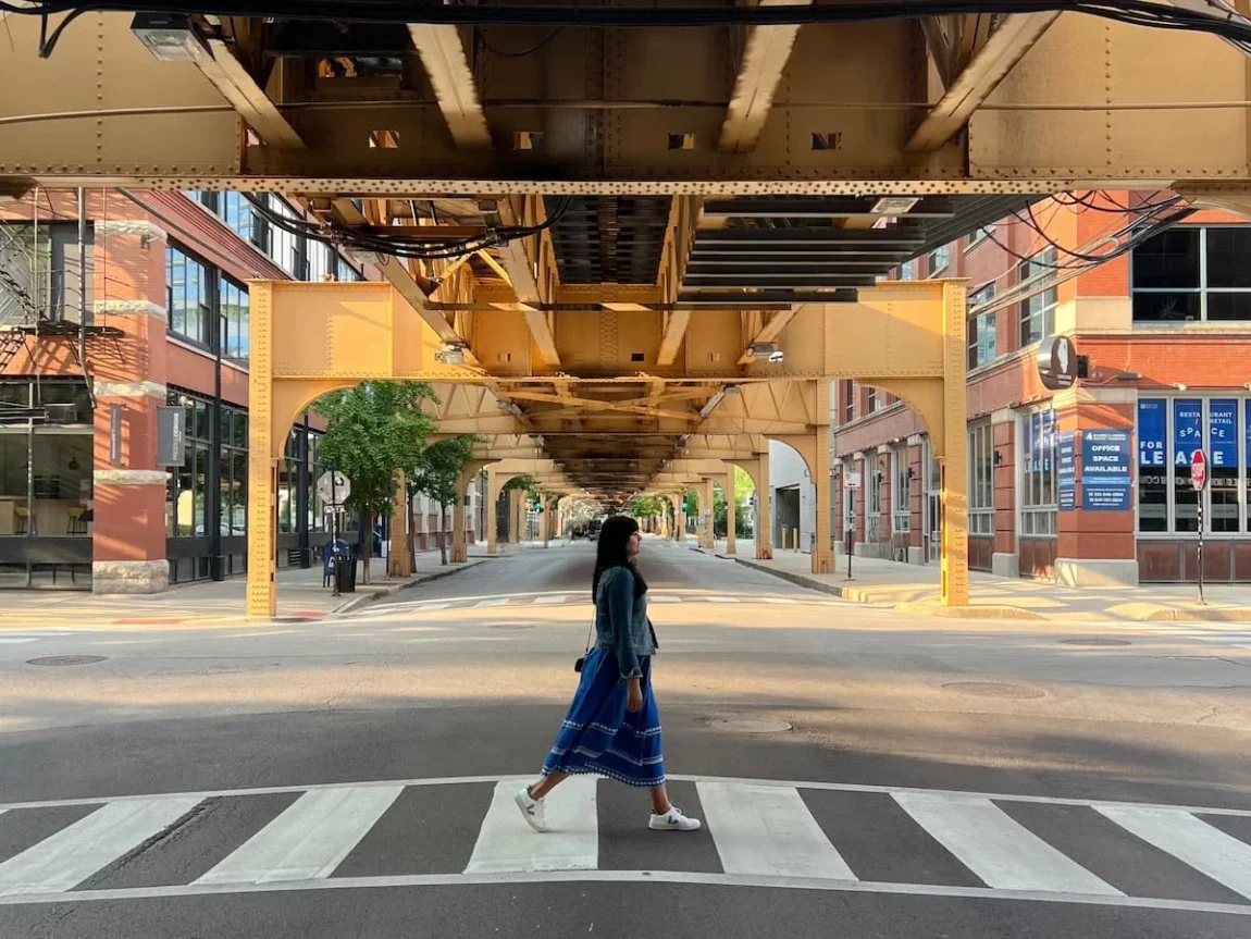 Bejal walking down North River road with the L train brides above, Chicago. Bejal is wearing a bright blue skirt and denim jacket with black cross body bag. She has white Veja trainers on.